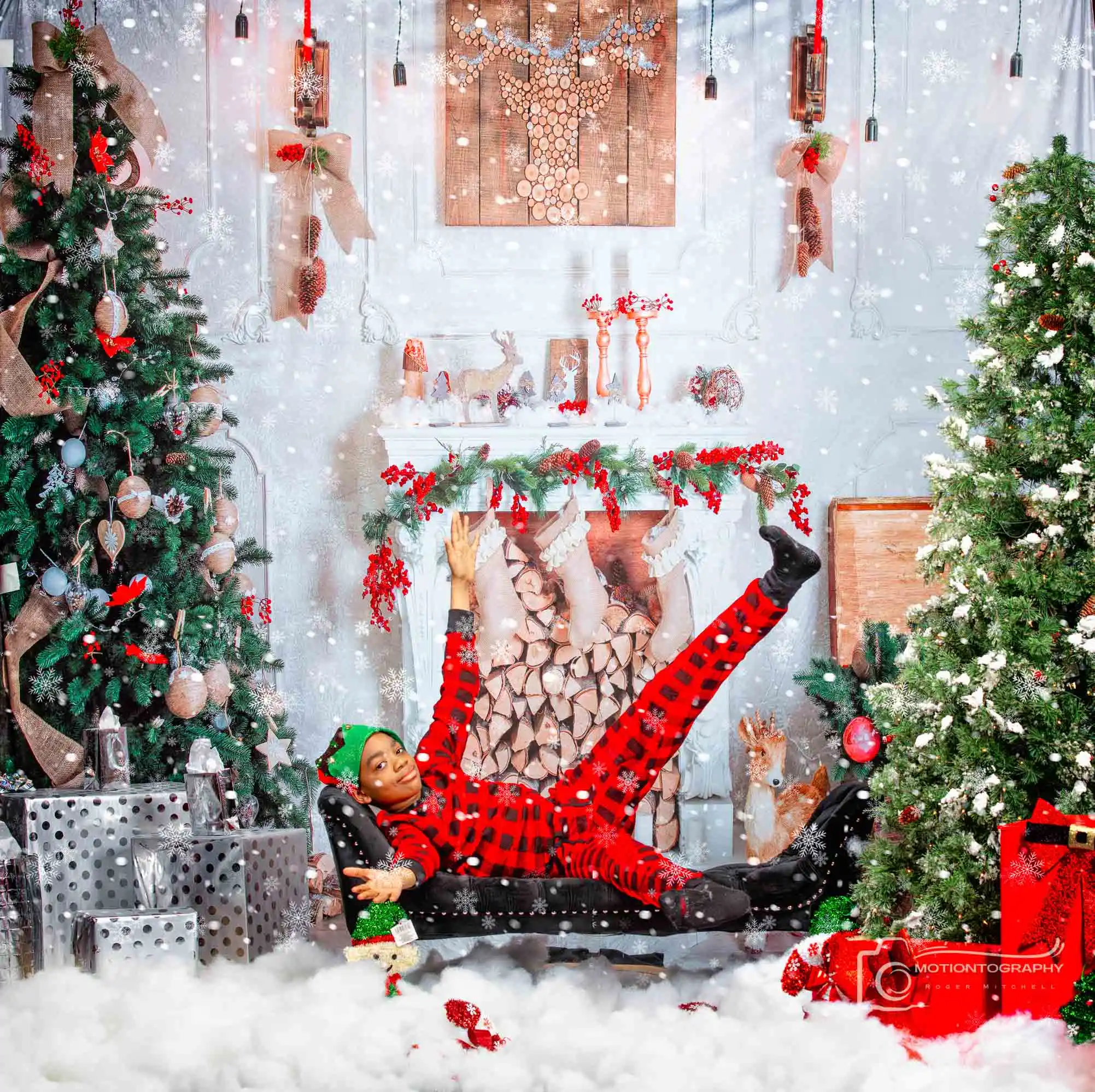 Playful Christmas portrait of a child in red plaid pajamas kicking their legs up on a chaise lounge amidst falling snow and holiday decor.