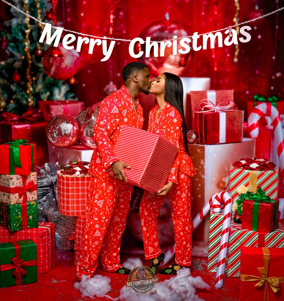 Couple in matching red Christmas pajamas kissing while holding a large gift box surrounded by piles of presents and holiday decor
