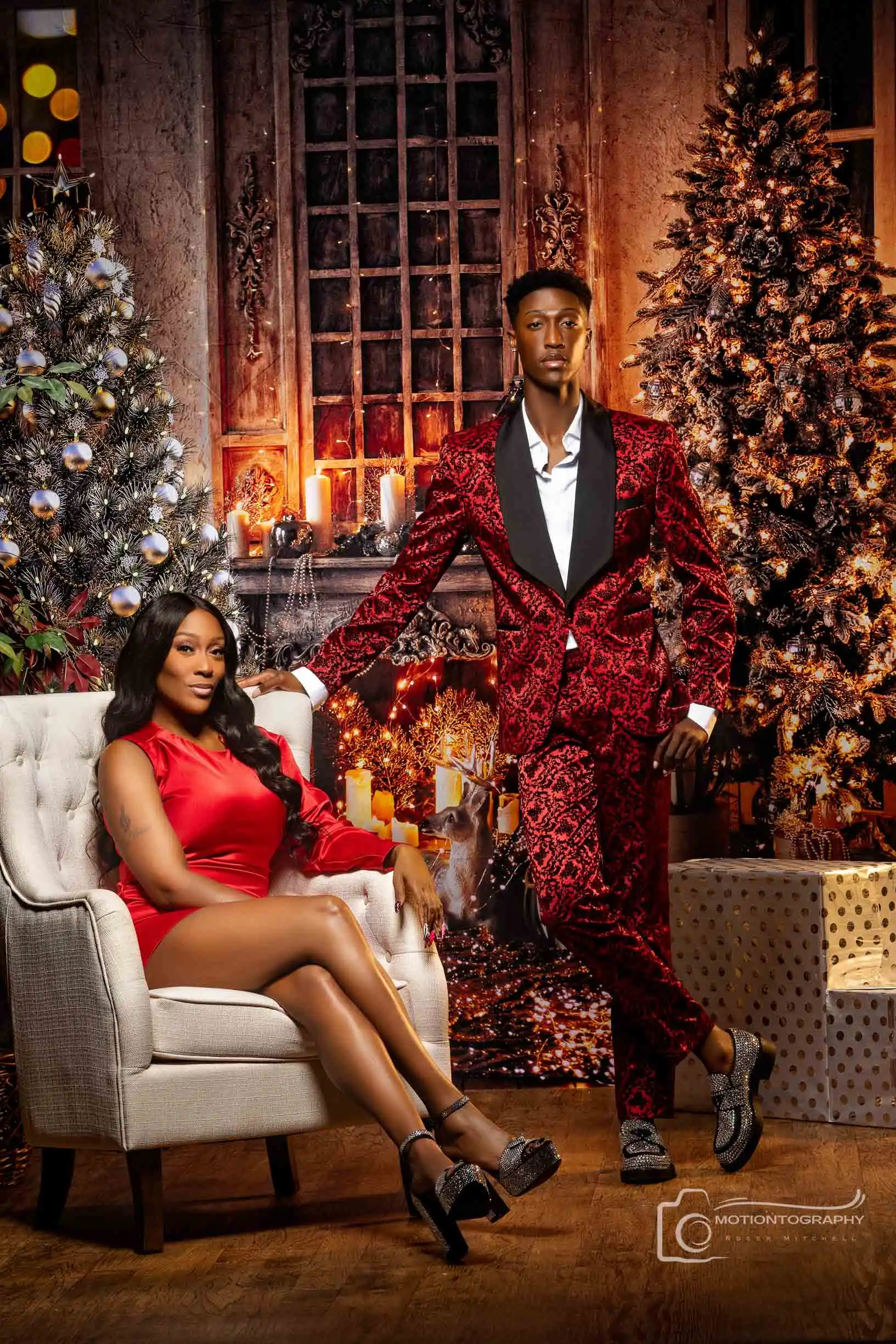 Elegant Christmas portrait of a mother in a red dress seated in a white chair and son in a patterned red suit, surrounded by festive holiday decor.