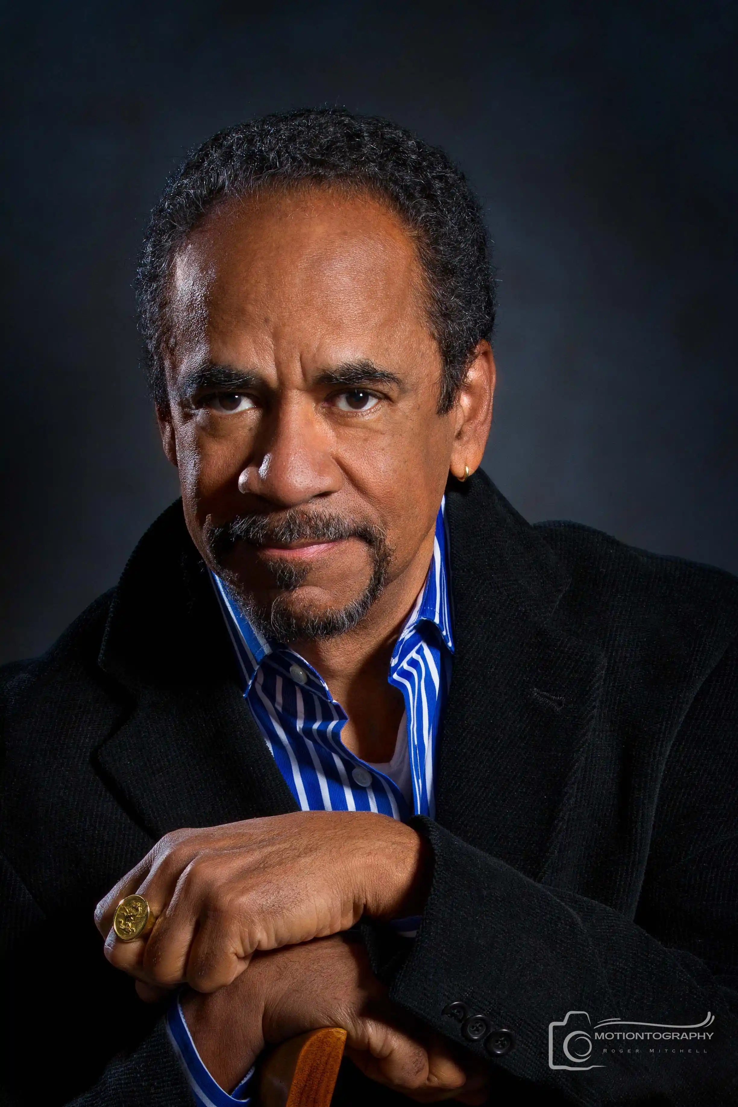 Tim Reid wearing a striped shirt and black blazer, posing with a serious expression against a dark background