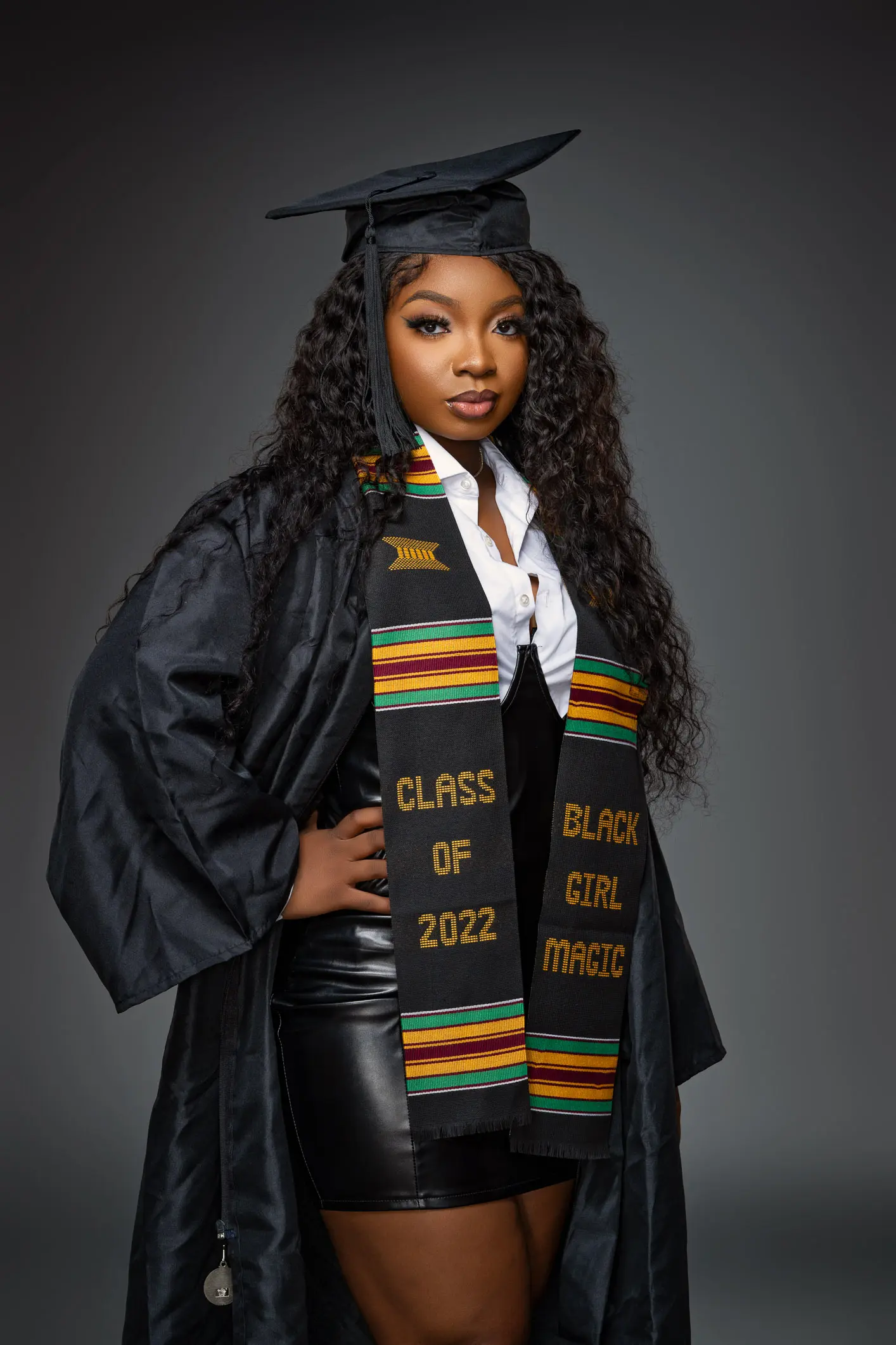 Studio graduation portrait of a woman wearing a black gown and a kente-style stole reading 'Black Girl Magic' and 'Class of 2022'.