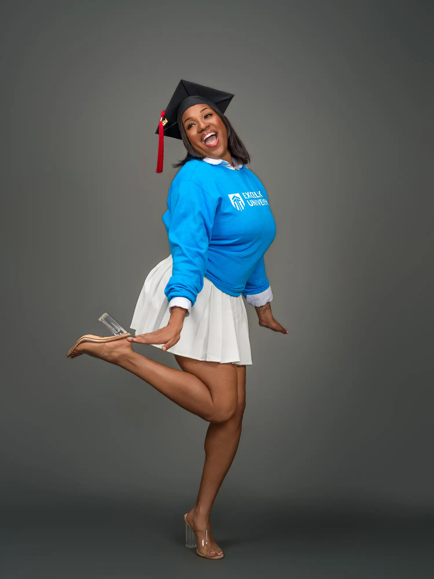 Energetic studio graduation portrait of an Excelsior University graduate jumping for joy in a blue sweatshirt and white skirt.