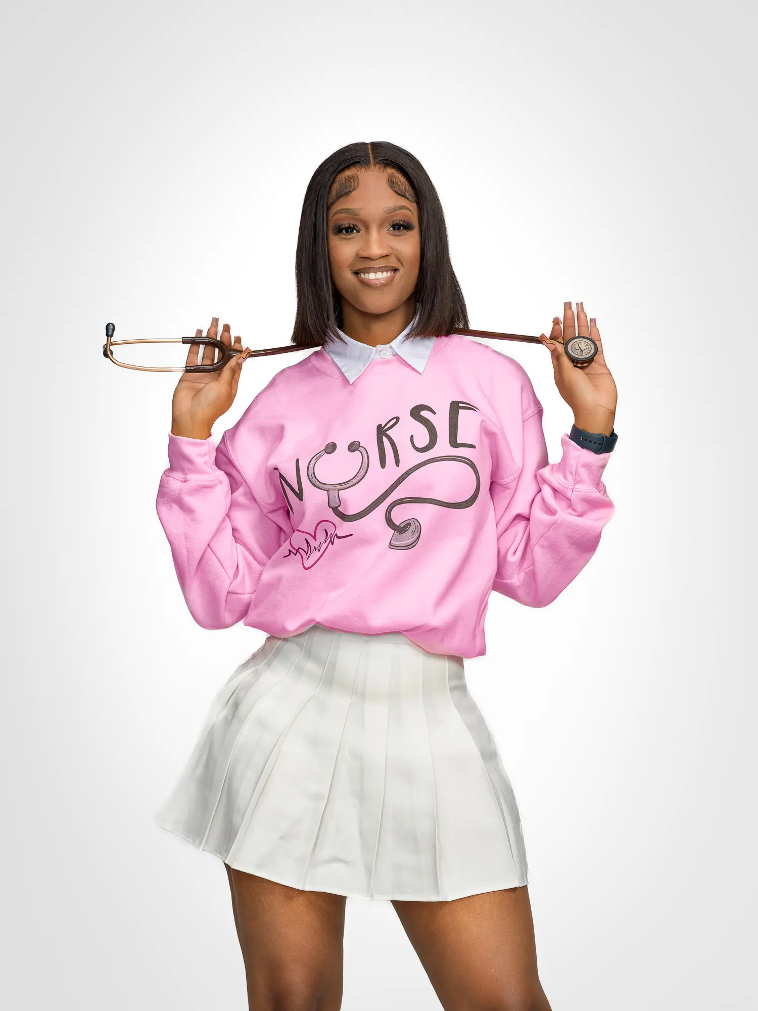 Studio graduation portrait of a nursing student wearing a pink nurse sweatshirt and white skirt holding a stethoscope.