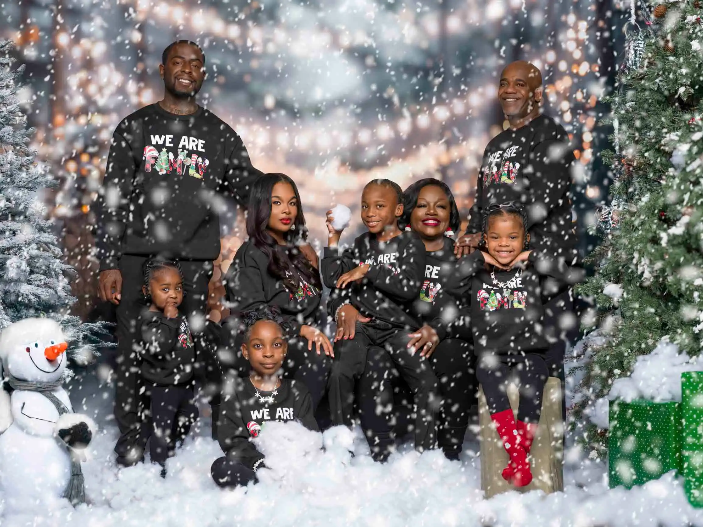 Large family in matching black WE ARE Christmas sweaters in fake snow with trees and snowman